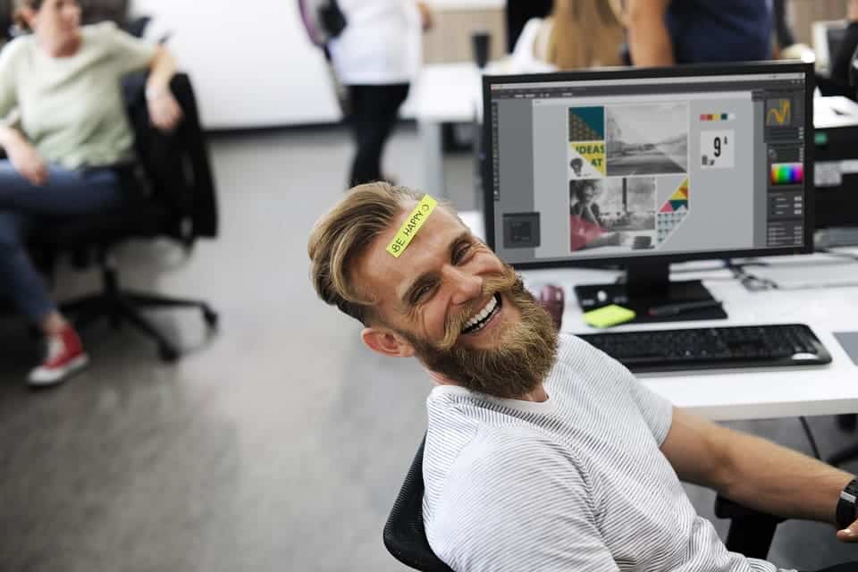 homem em um escritorio olhando para a camera rindo com um papel escrito be happy na testa